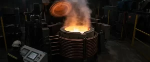 Operador com capacete branco e uniforme operando painel de controle em frente a uma fornalha de indução com metal fundido brilhante e vapor subindo, em ambiente industrial escuro com equipamentos e cercas de segurança ao fundo.